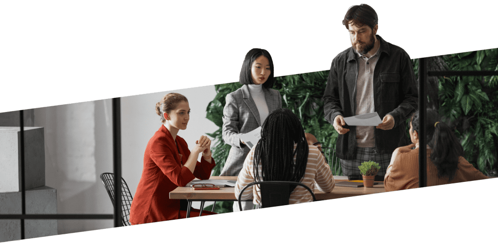 Professional meeting in a bright, modern workspace. Five staff members are engaged in a discussion around a table. The environment is enhanced by lush greenery in the background, adding a refreshing touch to the corporate setting.