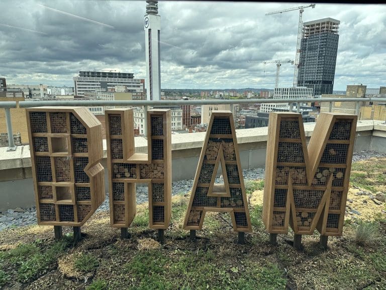 Large freestanding pollinator habitat letters spelling "bham" on a rooftop garden, each letter constructed from wooden frames filled with drilled logs, bamboo canes, and natural materials.