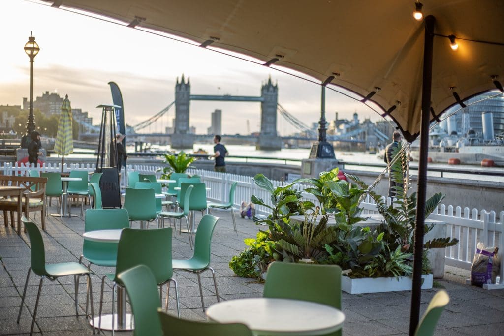 Outdoor seating under canopy with green tables and river view.
