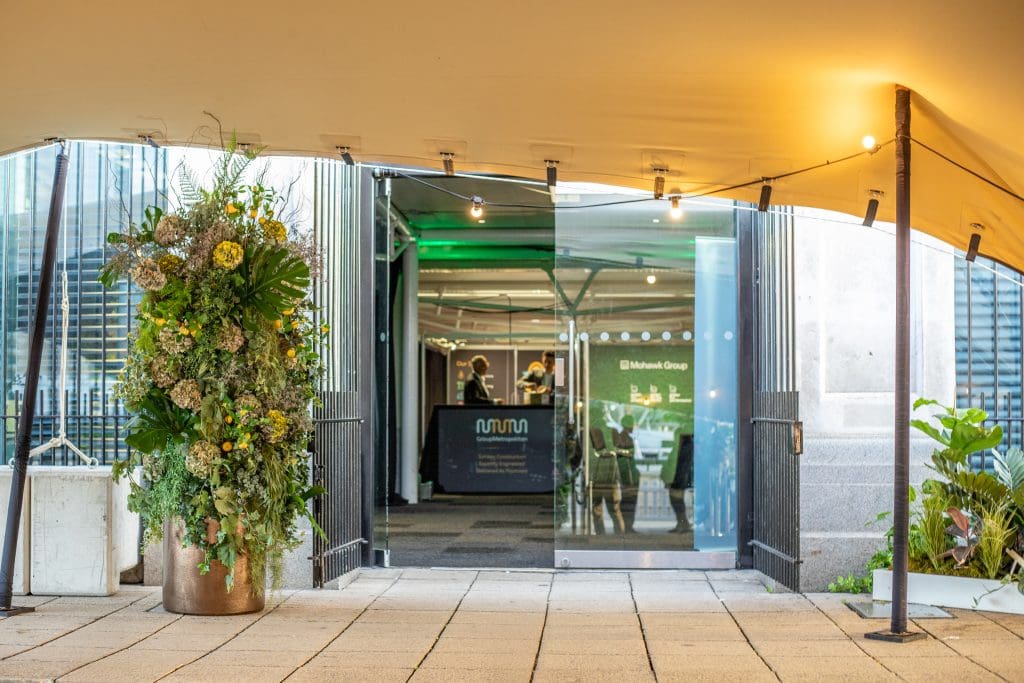 Event entrance with tall plant arrangements and illuminated cbre stand.