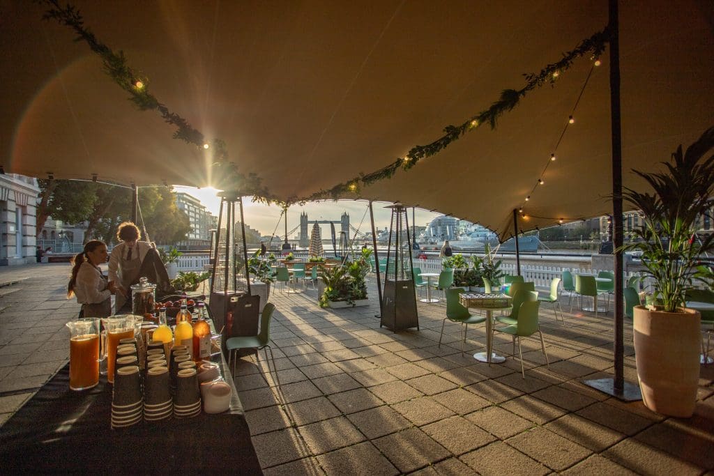Event dining setup with long table under marquee decorated with greenery and lights.