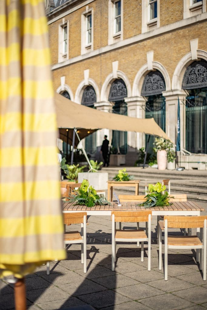 Cbre terrace with yellow-striped parasol, tables, and potted plants beside historic old billingsgate arches.