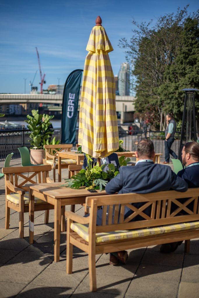 Outdoor seating area with wooden tables, striped umbrellas, and greenery at cbre event.