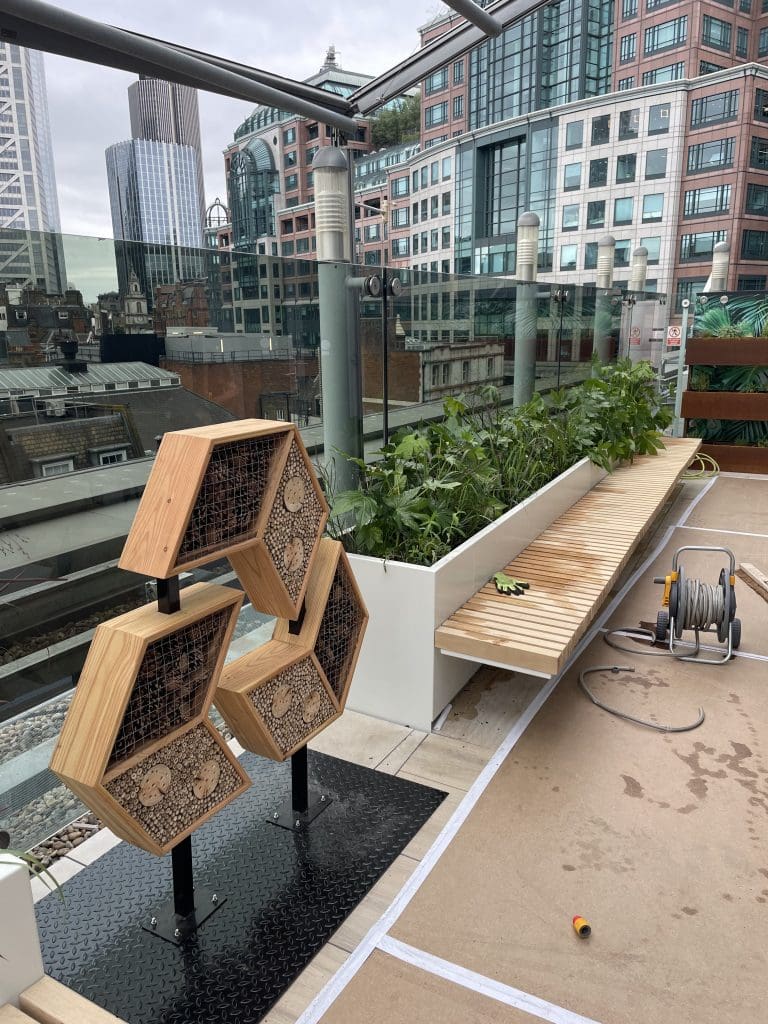 Rooftop terrace with stacked hexagonal pollinator habitats mounted on metal poles, overlooking modern office buildings.