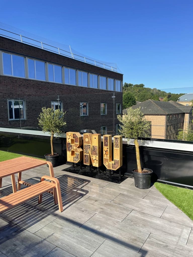 Rooftop garden installation with large wooden pollinator habitat letters spelling "bnu," raised above planters and filled with bamboo, logs, and cut wood.