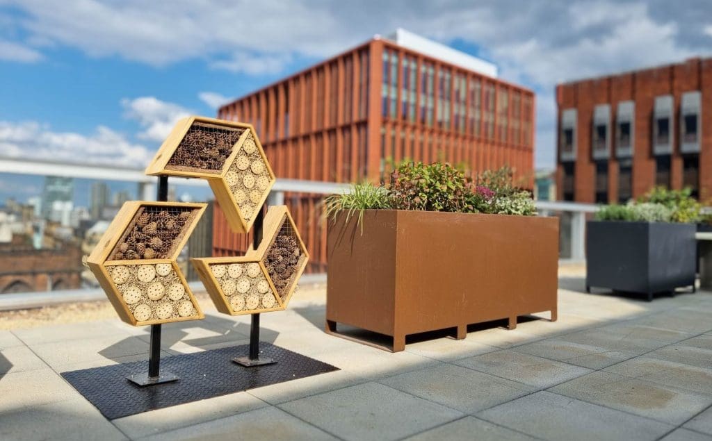 Rooftop pollinator habitat installation featuring stacked hexagonal frames filled with bamboo and wood, next to a red modern building.