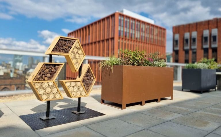 Rooftop pollinator habitat installation featuring stacked hexagonal frames filled with bamboo and wood, next to a red modern building.